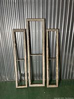 Three vertical rectangular wooden window frames with original paint finish, two smaller and one larger, displayed against a corrugated metal backdrop.