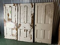 Three solid wood panel doors with peeling white paint, one showing natural wood underneath, standing vertically side by side.