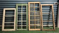 Four wooden window frames leaning against a metal wall, showing their size variation and paint condition.
