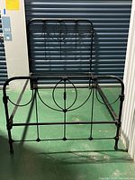 Full view of antique iron bed frame showing headboard, footboard, and bedrails, standing in a storage unit with a green floor and blue door.