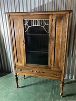 Front view of the antique wood curio cabinet showing glass door with decorative trim, turned legs, and bottom drawer.