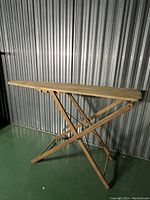 Side profile angle of antique wooden ironing board showing long rectangular top and foldable wooden legs with cross supports.