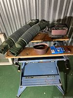 Black & Decker Workmate Model 400 portable workbench with wooden clamping tops and blue metal frame, black Black & Decker belt sander with sanding belt attached, extra sanding belts in original packaging, and two green accordion-style downspout extensions.