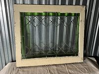 Front view of the rectangular stained glass window with green border, clear textured glass panes arranged in diamond pattern with lead cames, in off-white painted wooden frame.