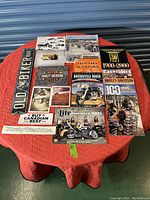Overview of the entire Harley-Davidson collectible lot showing magazines, posters, brochures, sticker, and tin sign on a red tablecloth.