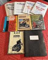 Collection of vintage motorcycle manuals and catalogs arranged on a red fabric surface, showing titles and covers.