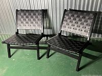 Two black woven faux leather mid-century modern style chairs with dark wood frames, shown from front on a green floor with metal wall background.