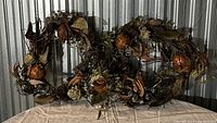 Pair of custom wreaths made with faux pomegranates, artichokes, pinecones, dried leaves, and various faux natural botanical elements displayed against a metal corrugated backdrop on a cloth-covered table.