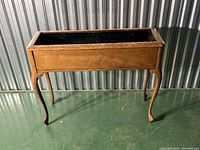 Wood planter with Queen Anne style legs, showing front and side profile, placed on green floor against corrugated metal wall.