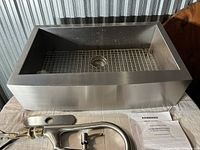Apron-front stainless steel farmhouse sink with bottom grid, drain plug, faucet components, and installation guides.