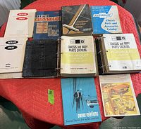 Collection of 11 vintage General Motors manuals and catalogs spread on a red surface, showing the different volumes and binders including Chevrolet service and overhaul manuals and parts catalogs.