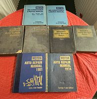 Photo showing all nine manuals arranged on a red surface: two Motor Air Conditioner & Heater Manuals in light blue softcovers, seven Motor's Auto Repair Manuals in black and blue hardcover editions from years 1962 through 1981.