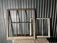 Two wooden window frames leaning against metal wall, one large no glass, one medium with broken panes