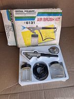 Top-down view of airbrush kit in original white molded tray inside box, showing airbrush, hose, two jars, and accessory.