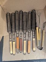 Seven black metal BBQ grilling baskets of various wooden handle colors and two wooden handle grilling tools inside a cardboard box