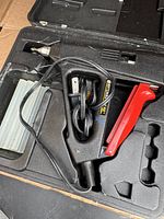 Open black molded plastic case showing electric hot glue gun with red trigger, power cord, and several clear glue sticks.