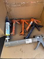 Top view of six caulking guns in a cardboard box, showing their colors and brand markings.