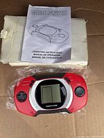 Front view of red handheld electronic game console with circular screen and included operating manual on top of the box.