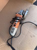 Orange and silver Black & Decker electric angle grinder laid on cardboard, showing tool body, power cord wrapped, and blade guard.