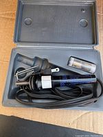 Soldering iron kit inside grey plastic carrying case. Iron with attached power cord, metal stand on iron, and small plastic container with cleaning sponge or wire.
