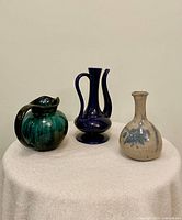Three mid-century pottery pieces placed on a light cloth-covered surface against a plain background: a green and dark blue pitcher, deep blue double-handled vase, and cream and blue small vase.