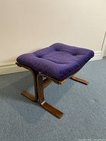 Front view of mid-century modern ottoman with purple tufted velour cushion and bent plywood frame on carpeted floor against beige wall.