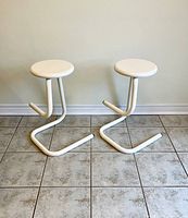 Front view of both white tubular steel stools showing round seats and paperclip-shaped frames