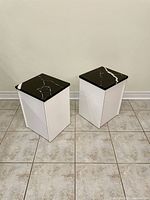 Two tall rectangular pedestals with white melamine bodies and black quartz tops marked with white veining. Positioned on tile floor against a plain wall.
