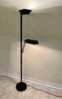 Full view of black floor lamp showing tall pole, circular base, upward facing top light, and articulating lower light.
