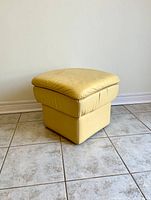 View of pale yellow leather and vinyl ottoman showing its top and side profile on tiled floor.