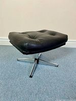 Front side angle of the ottoman showing the padded vinyl seat and chrome swivel base with four legs.