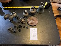 Full collection of assorted small vintage collectibles on black surface with measuring tape showing scale. Includes bells, Buddha figurines, animal figures, and medallion.