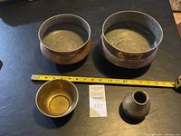 Four metal bowls and one small metal vase on a dark surface with a tape measure for scale. Two large copper bowls about 10 inches wide.