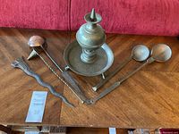 Full view of all items: brass incense burner, wavy keris part, copper ladle, two brass spoons on wood table against red couch