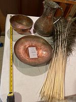 View of entire lot on tabletop featuring three copper items and peacock feathers with measuring tape for scale