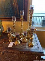 Seven brass candle holders of various designs and heights with purple candles in glass votives on an ornately carved wooden chest by a window overlooking water.