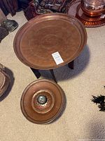 Copper tray table on carved wooden folding stand with separate copper item beside it on carpet.