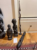 Four carved African wooden statues displayed on a wooden floor near a wall and cabinet. Various styles including tall abstract woman, seated man, wooden laying figure, and standing figure with woven cover.