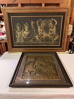 Both Thai framed artworks on display, one leaning against the wall and one flat on table