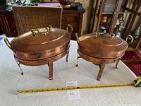 Two copper chafing dishes side by side on a table with measuring tape showing size, oval shape, with lids and brass handles on stands.