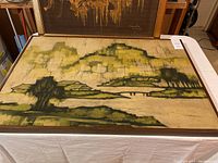 Photo of smaller framed batik artwork with a landscape scene in green and yellow tones.