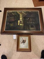 Two paintings side by side on the floor showing full view. Larger village scene painting and smaller portrait painting below it.