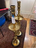 Two tall brass candle holders with wide circular trays and wooden bases shown side by side on a carpeted floor near furniture.