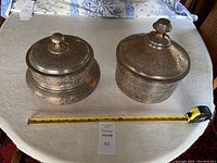 Two copper lidded pots side by side on table with tape measure in front