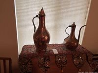 Photo showing both large vintage copper Turkish pitchers with detailed patina and engraving, placed on a patterned fabric surface beneath window blinds.