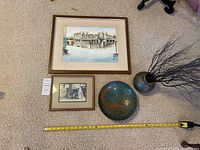 Overview of all items on carpet including the 2 framed street scene artworks, brass and enamel round dish, brass urn with black decorative branches, and yellow measuring tape indicating size.