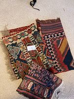 Three vintage flat weave rugs folded and laid on carpet, showing patterns and fringes.