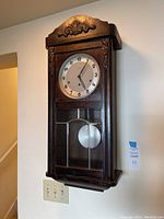 Full view of the dark wooden wall clock mounted on a wall, showing the carved decorative top and glass pendulum door.