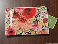 Front view of the Kate Spade floral pencil case with tag attached, displaying the colorful floral pattern and gold zipper.