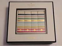 Top-down view of the four erasers in a white box with black trim and Kate Spade branding in gold, showing the multicoloured horizontal stripes and gold spade logo on each eraser.